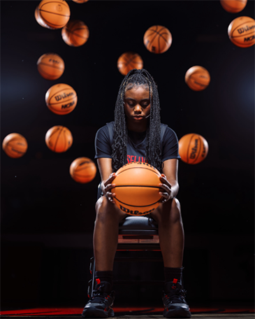 A high school basketball player sitting in a chair in a dark gym. She is staring down at a basketball in her hands as multiple basketballs fall in the background behind her.