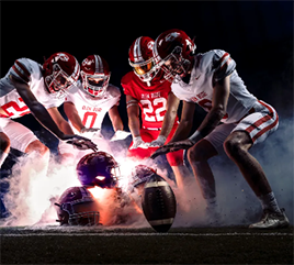 Four high school football players on a dark field. They are looking at and holding their hands out to a pile of helmets and footballs, as if warming their hands by a campfire, with fog all around their legs.