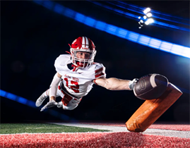 A high school football player jumping towards the camera and reaching forward with a football in his hand.