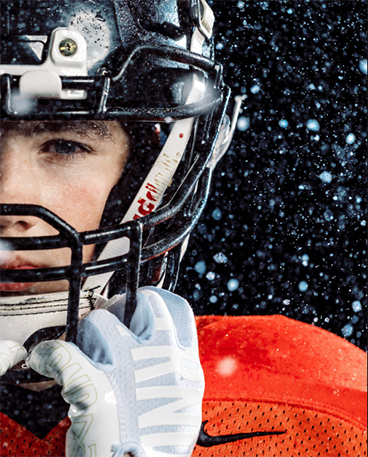 A closeup portrait of a high school football player's face and shoulders. Only the left side of him is in the frame. He is looking at the camera with his helmet and uniform on, and there are water droplets in the air around him.