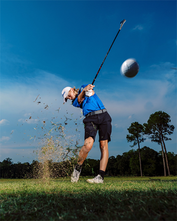 A high school golf player with her golf club mid-swing. There is a golf ball in the foreground, mid-air, with grass around it.