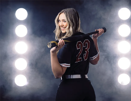 A high school softball player, standing in front of stadium lights peeking through fog. She is posing with her back to the camera, and her bat across her shoulders. Her head is turned towards the camera and she is smiling.