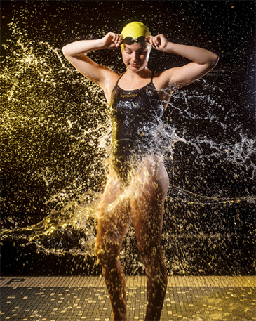 A high school swimmer standing in her swimsuit, with her goggles on her head. She is looking at the ground and smiling as she places her hands on her goggles and water is splashing all around her.