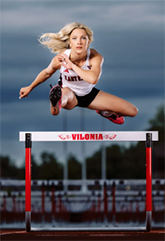 A high school track-and-field athlete jumping over a hurdle. She is facing straight towards the camera and is mid-jump over the hurdle.