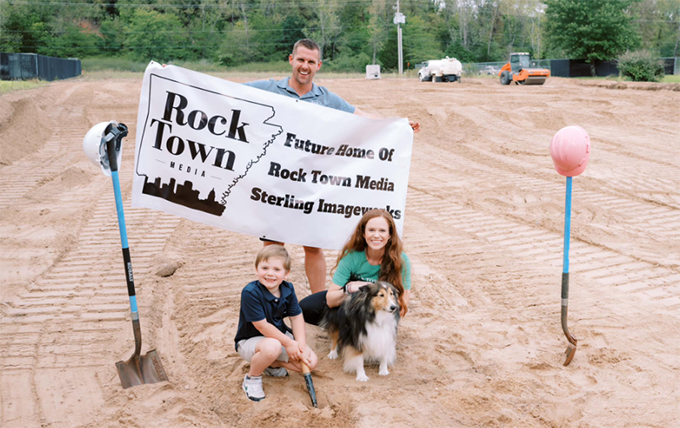 Jaison, Callie, and Elias Sterling posing with their dog in front of a dirt lot with construction vehicles in the background. Jaison is holding a banner that reads: Future Home of Rock Town Media Sterling Imageworks