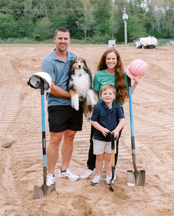 Jaison, Callie, and Elias Sterling posing with their dog in front of a dirt lot with construction vehicles in the background.