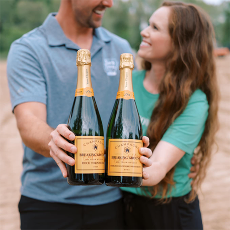 Callie and Jaison Sterling holding smiling at each other and holding two bottles of champagne. The labels on the bottles read, Breaking ground, congratulations on your studio